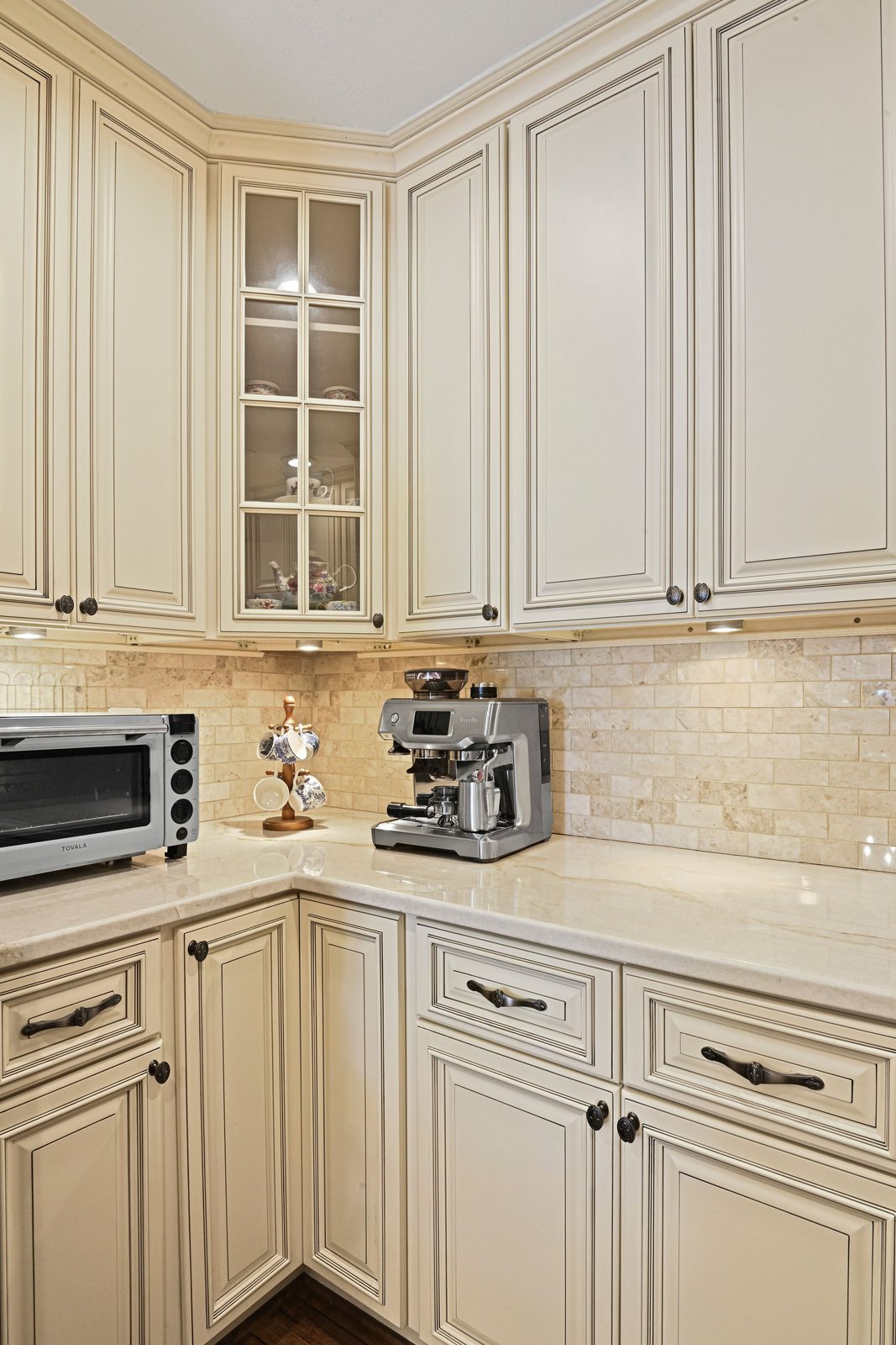 Corner of cream cabinetry with glass-front door, stone counters, tile backsplash
