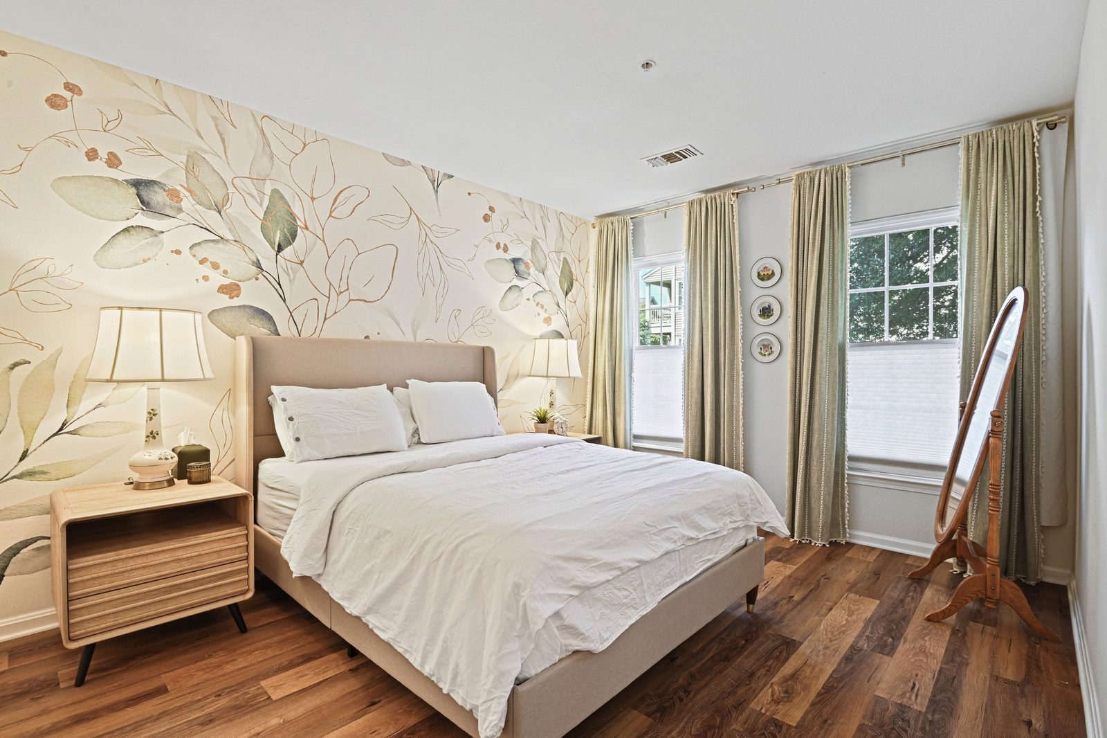 Bedroom with botanical mural accent wall, two curtained windows and wood-look floors