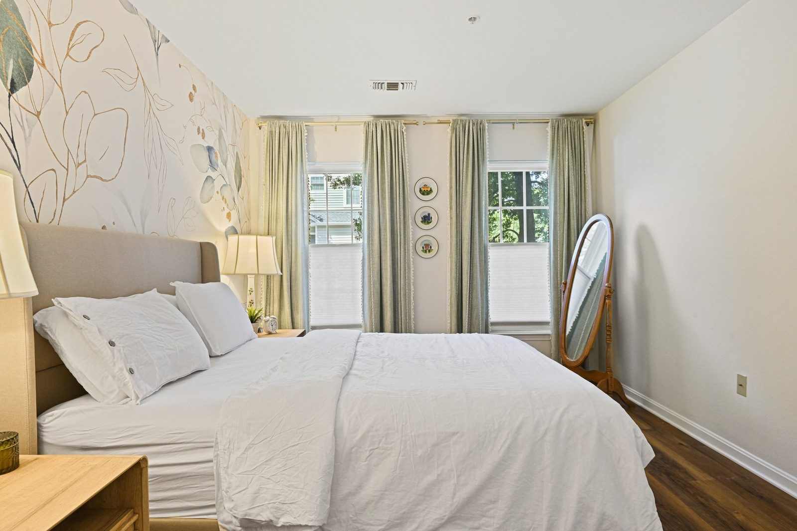 Bedroom with two curtained windows, cheval mirror, mural wall and wood-look floors