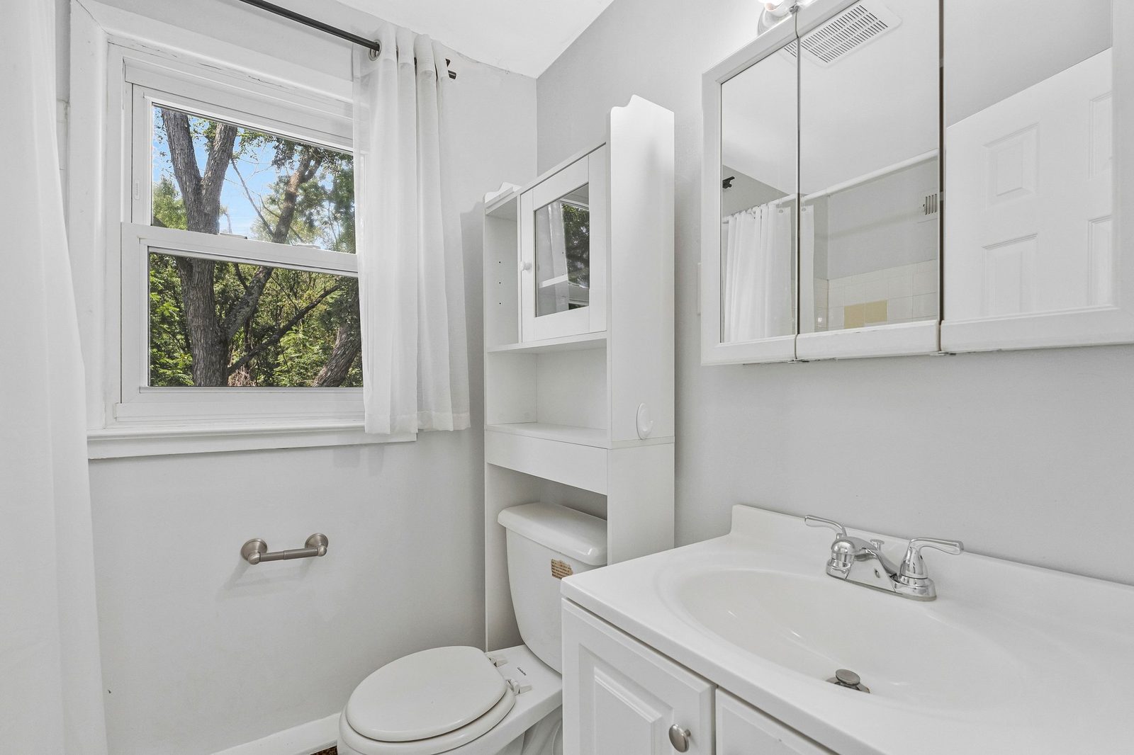 White bathroom with single vanity, mirrored cabinet and over-toilet shelving
